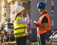 Two construction workers in hard hats and vests, one gesturing toward building, the other looking