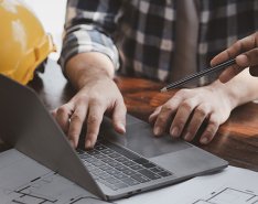 Person using laptop, with another person gesturing with a pencil. Yellow hard hat is beside them