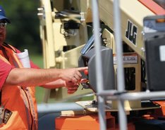 A construction worker in a bright safety vest and blue baseball hat presses a button on a piece of machinery