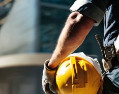 Person in gloves and vest holding yellow hard hat in front of building