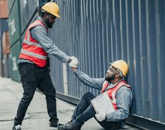 Two workers in vests and hard hats; one is being helped up by the other