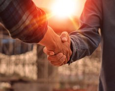 Two people shaking hands; one person holds blueprint; sun shines above handshake