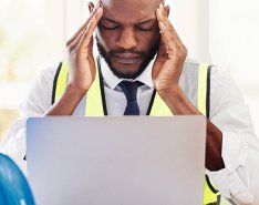 Man wearing vest looking at laptop and holding head in frustration with hardhat on desk