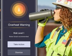 A woman with long curly hair, wearing a hard hat, a bright safety vest, sunglasses and gloves drinks from a water bottle. A phone screen can be seen displaying a temperature of one hundred degrees and text saying "overheat warning: risk level 1"