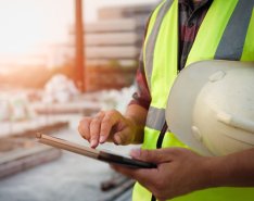 A construction worker wearing a bright safety vest has a hard hat tucked under their arm and is checking a tablet