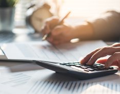 Two hands on a desk; one hand holding pencil, the other using calculator