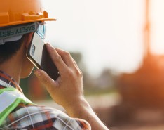 A person wearing an orange hardhat and a bright green safety vest, facing away from the camera, is holding a cellphone to their ear.