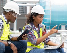 Two workers in vests and hard hats. Both looking at computer and talking