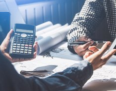 Two people sitting at desk one; one holds calculator, the other holds a pen and tablet