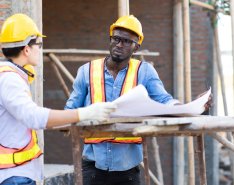 Two people wearing hard hats and bright safety vests talk over a large sheet over paper on a construction site