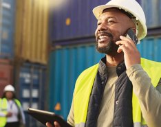 Worker in hard hat and vest using phone & smiling/Adobe Stock
