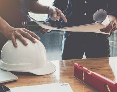 Two workers in office looking at plans; hard hats on desk 