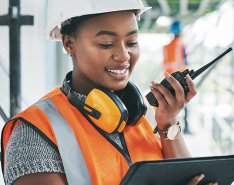 Woman with orange work vest, hard hat and headset looking at screen and holding comms device