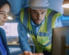 Woman in business attire on computer while man in vest and hard hat looks at screen