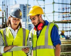 Three individuals in hard hats and bright safety vests look at a tablet display while standing on a construction site