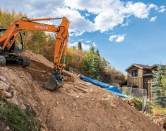 An excavator digs on the side of a hill with a house in the background