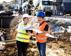 Two people in hard hats and bright safety vests are having a conversation while standing on a construction site