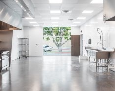 A bright room features an industrial work sink and two stovetops and ovens. A set of wire shelves sits in the back corner. A large window is at the back of the room near an open door. The floor is gray concrete and the walls are white.