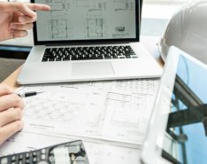 A laptop computer, tablet, papers, calculators, hardhat and three hands can be seen. One hand points to the computer screen while another holds a pen. Another hand points at the tablet screen.