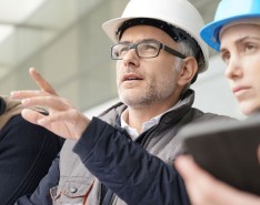 Two people wearing hardhats look forward. One of the people is wearing glasses and pointing at something out of frame. A third person, also wearing a hard hat, looks down.