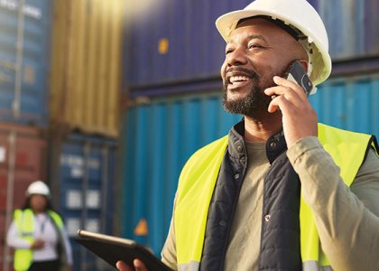 Worker in hard hat and vest using phone & smiling/Adobe Stock