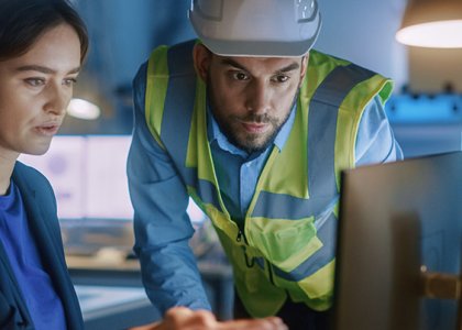 Woman in business attire on computer while man in vest and hard hat looks at screen